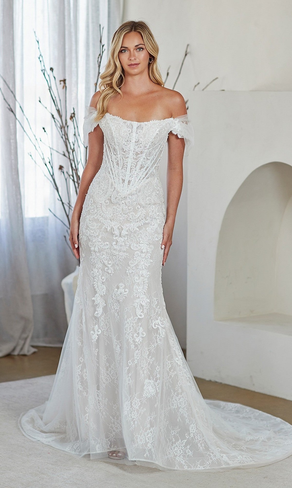 Woman in a white off-the-shoulder Amelia Couture 2153 long prom dress with lace details, fitted bodice, and slight train, standing indoors against a light background.